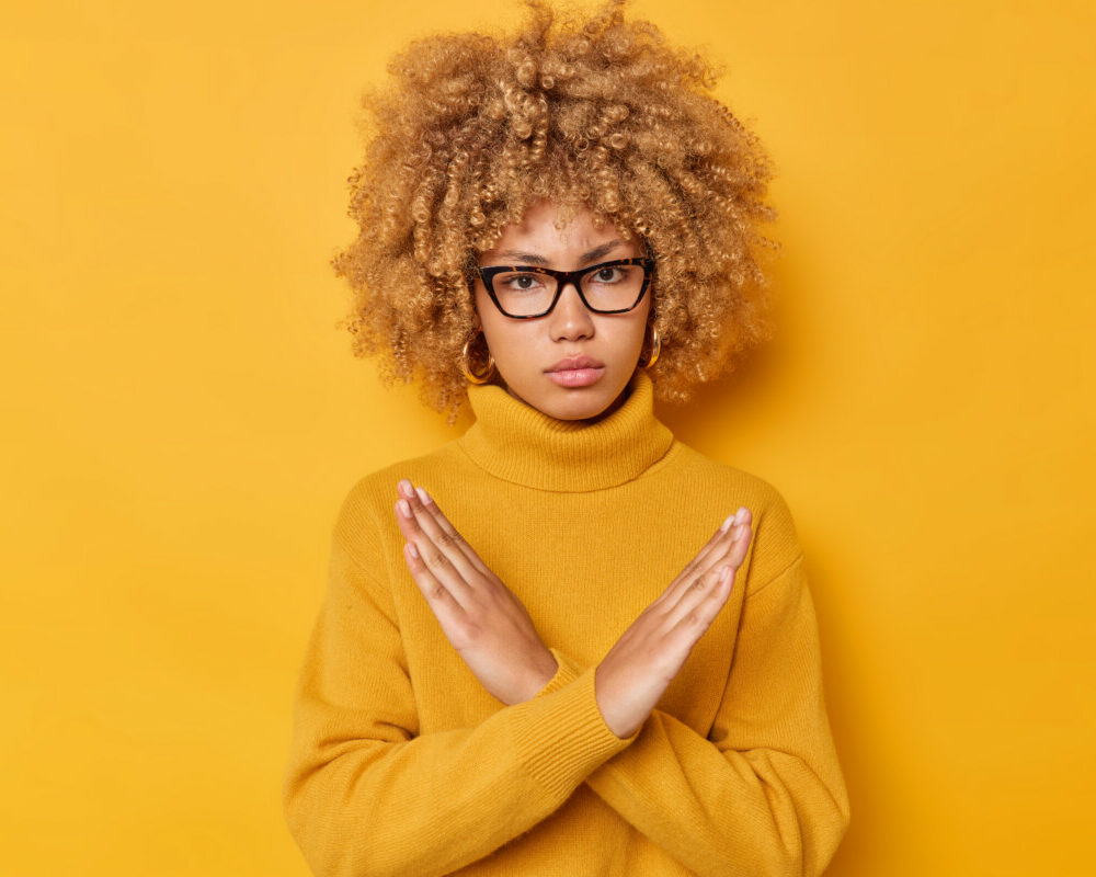 blond-curly-haired-woman-shows-cross-makes-stop-gesture-says-no-with-angry-expression-declines-something-wears-spectacles-long-sleeved-jumper-isolated-yellow-background-its-taboo