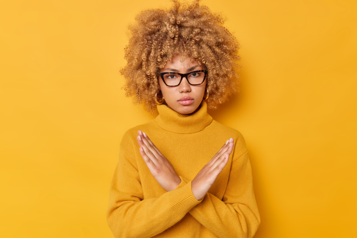 blond-curly-haired-woman-shows-cross-makes-stop-gesture-says-no-with-angry-expression-declines-something-wears-spectacles-long-sleeved-jumper-isolated-yellow-background-its-taboo