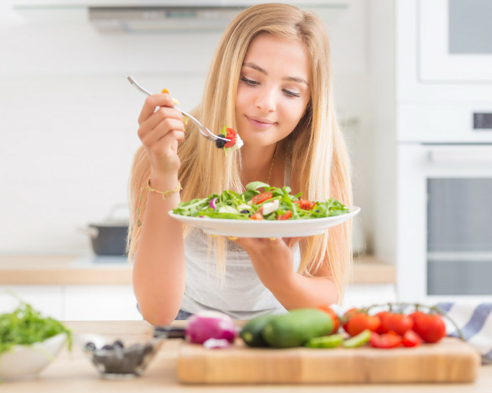 jeune-fille-blonde-heureuse-mangeant-salade-santé-d'araucula-d'épinards-de-tomates-d'olives-d'oignons-d'huile-d'olives