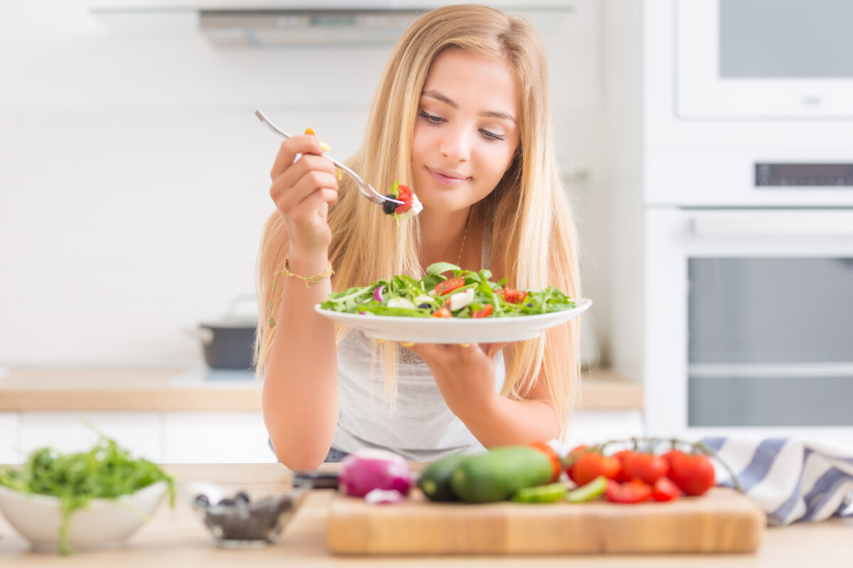 jeune-fille-blonde-heureuse-mangeant-salade-santé-d'araucula-d'épinards-de-tomates-d'olives-d'oignons-d'huile-d'olives