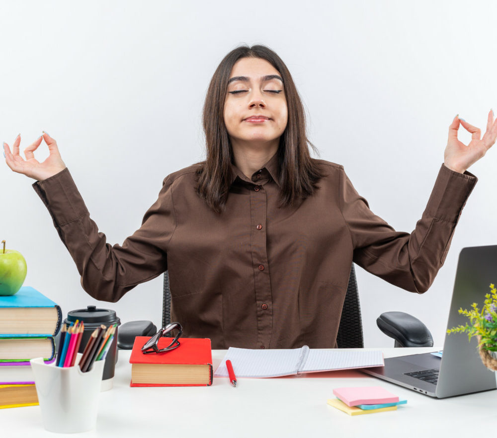 pleased-with-closed-eyes-young-school-woman-sits-table-with-school-tools-doing-meditation-gesture