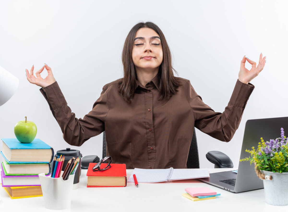 pleased-with-closed-eyes-young-school-woman-sits-table-with-school-tools-doing-meditation-gesture