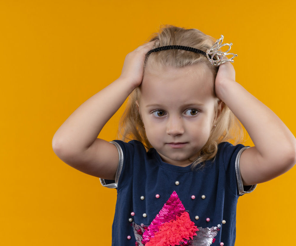 pretty-little-girl-wearing-navy-blue-shirt-crown-headband-holding-hands-her-head-looking-side-orange-wall