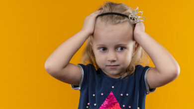 pretty-little-girl-wearing-navy-blue-shirt-crown-headband-holding-hands-her-head-looking-side-orange-wall