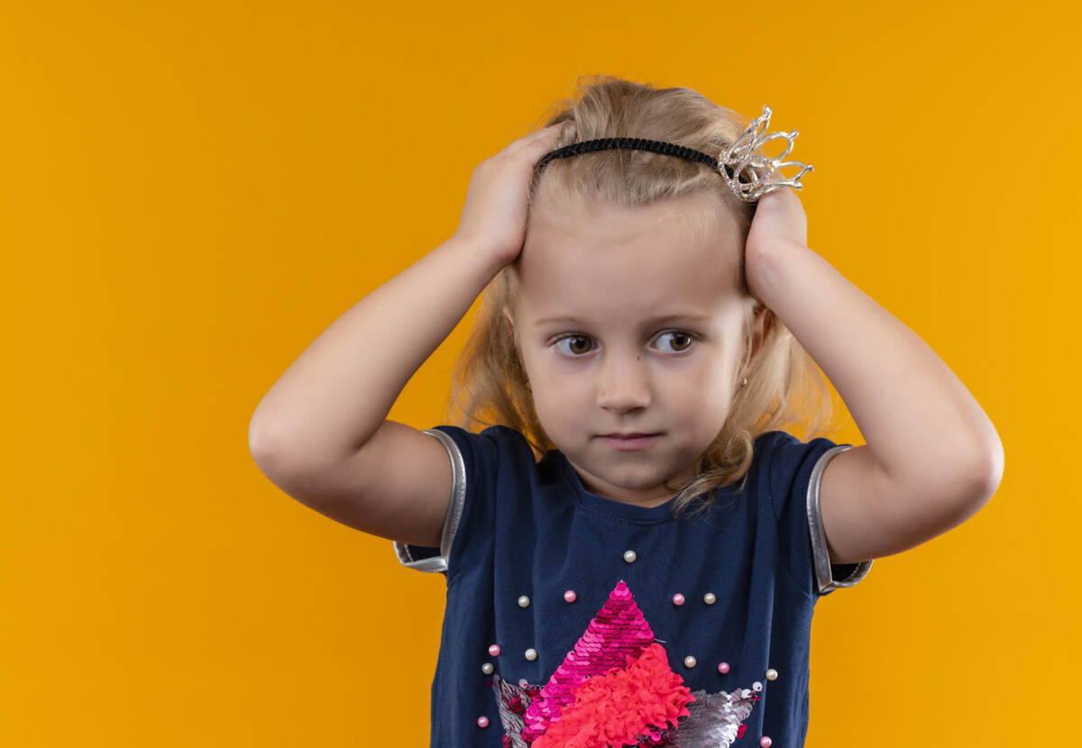 pretty-little-girl-wearing-navy-blue-shirt-crown-headband-holding-hands-her-head-looking-side-orange-wall