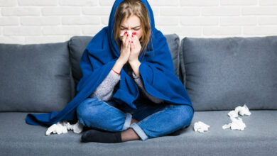 sick-woman-with-rheum-headache-holding-napkin-sitting-sofa-with-coverlet-pills-home