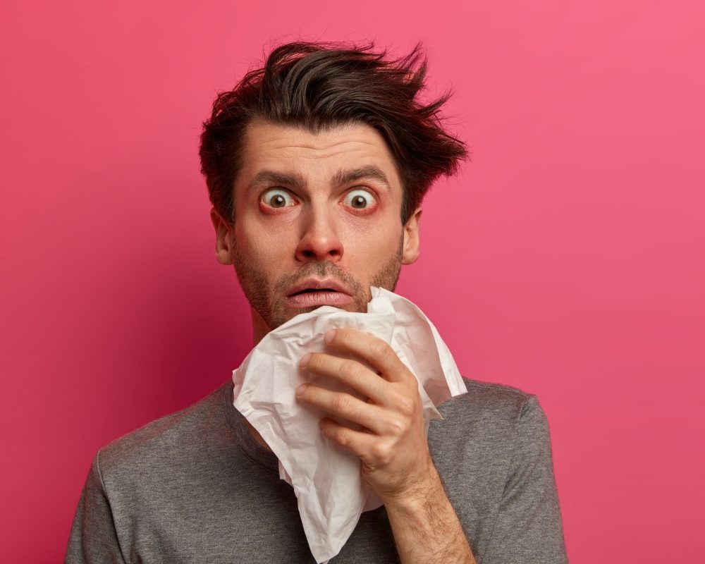 stunned-sick-man-has-flu-virus-allergy-respiratory-red-watery-eyes-blows-nose-tissue-finds-out-about-serious-disease-poses-pink-wall-health-medicine-symptoms-concept (1)-grippe