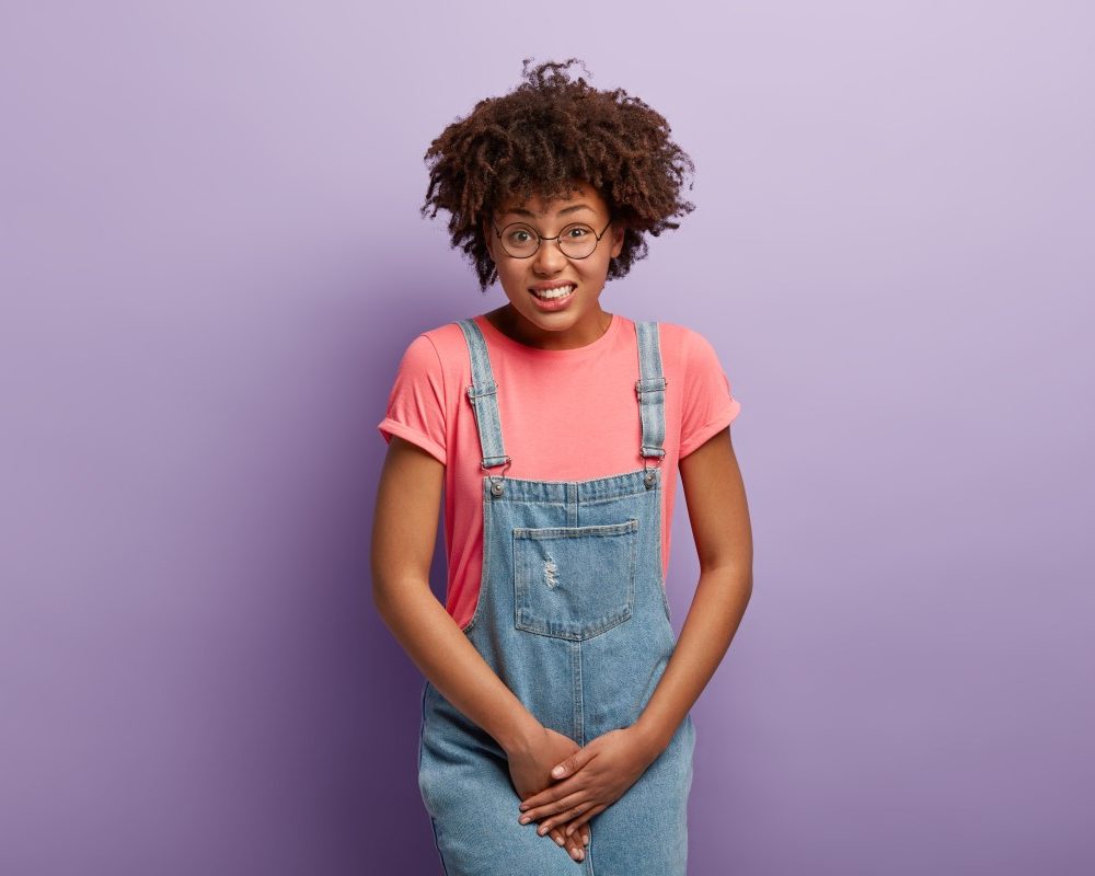 unhappy-dark-skinned-woman-holds-crotch-needs-toilet-has-problematic-situation-wears-pink-t-shirt-denim-sarafan-suffers-from-cystitis-isolated-purple-wall-people-urgency