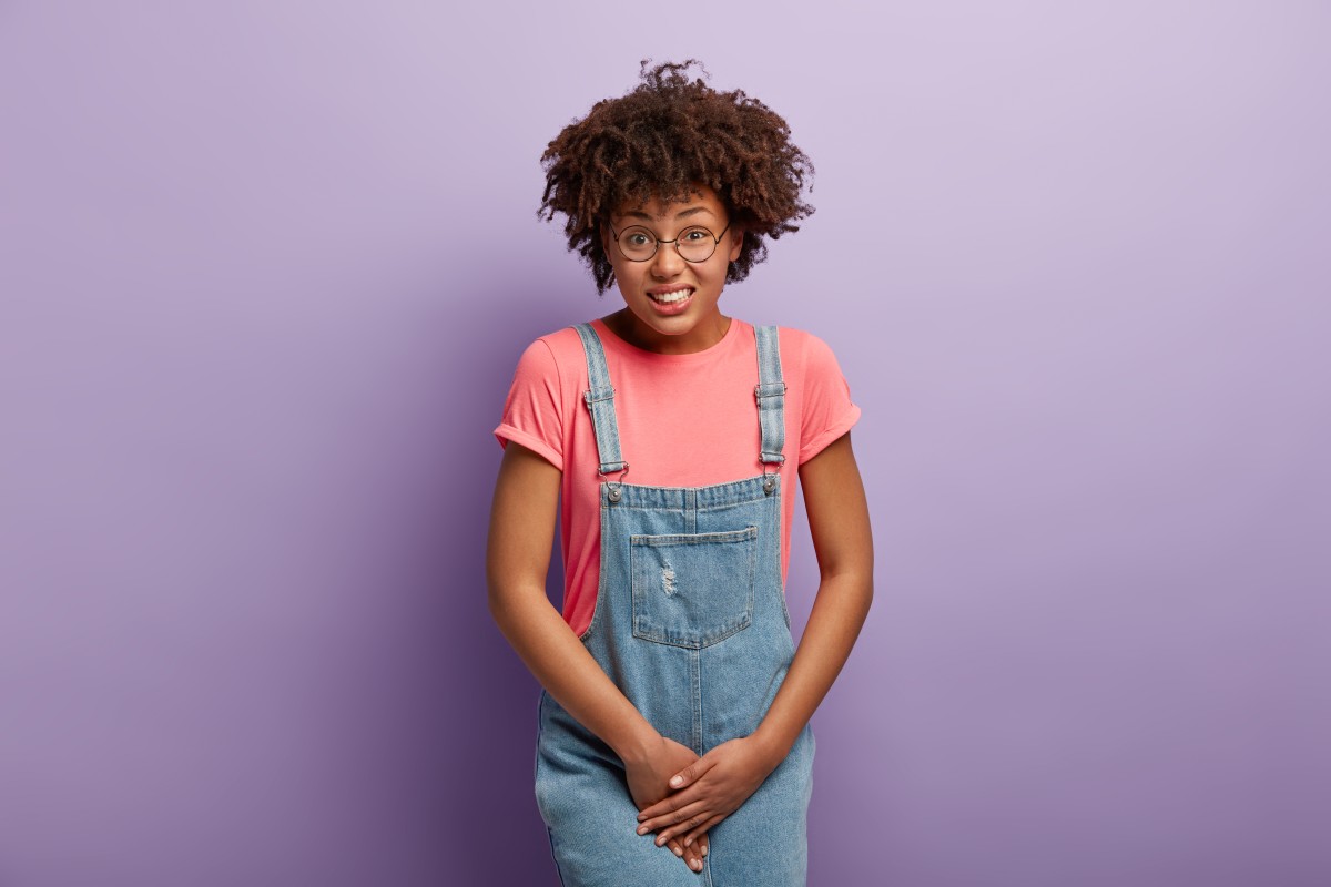 unhappy-dark-skinned-woman-holds-crotch-needs-toilet-has-problematic-situation-wears-pink-t-shirt-denim-sarafan-suffers-from-cystitis-isolated-purple-wall-people-urgency